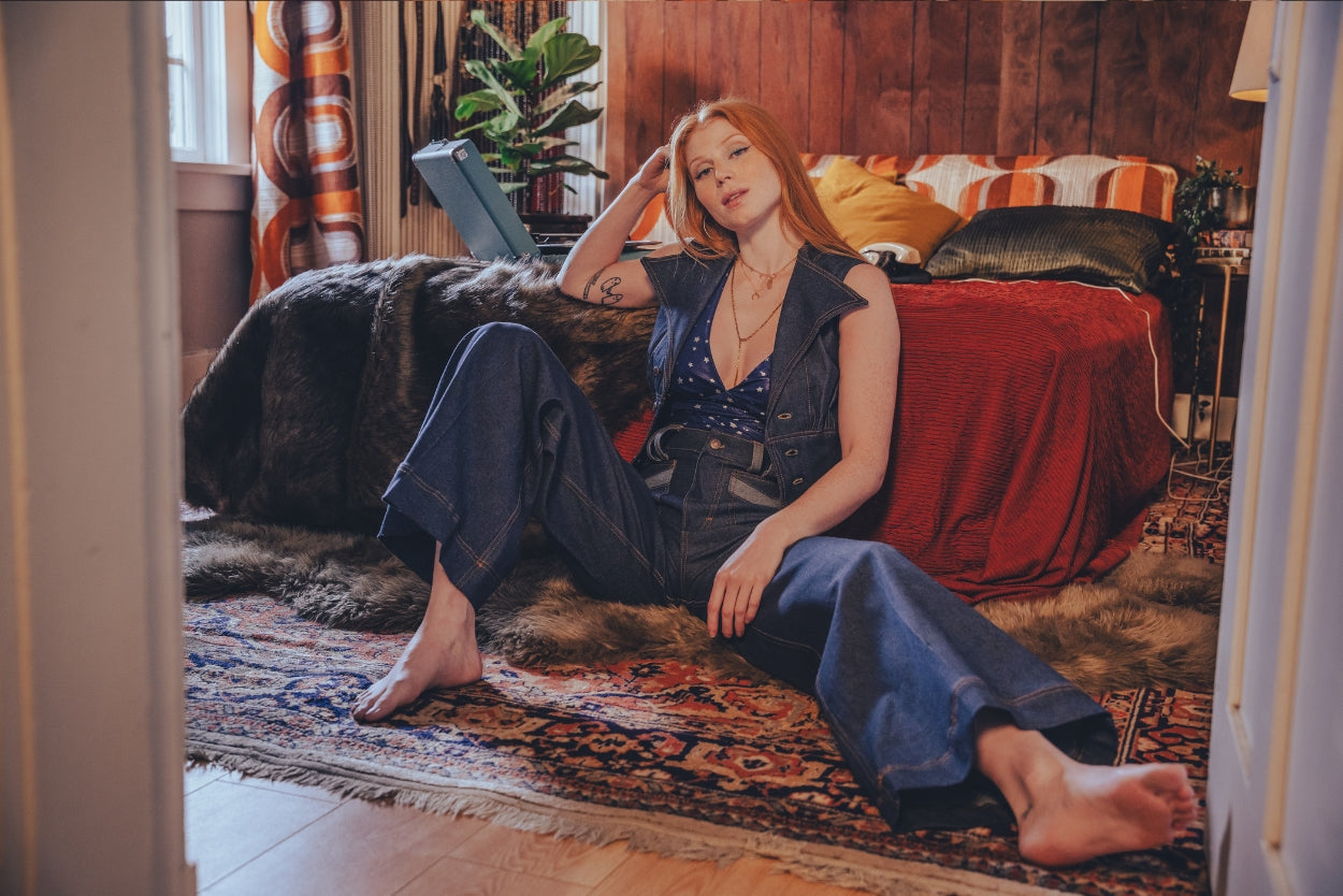 Girl sitting on the floor wearing a denim waistcoat and high-waisted 1970s-style bell-bottom jeans, showcasing retro 70s fashion, vintage-inspired denim, and sustainable small-batch style.