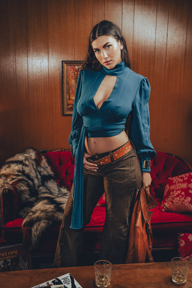 Gorgeous woman wearing a sexy teal peekaboo blouse with puffed long sleeves and a front tie paired with high waisted brown 70s leather jeans posed in a retro 1970s living room