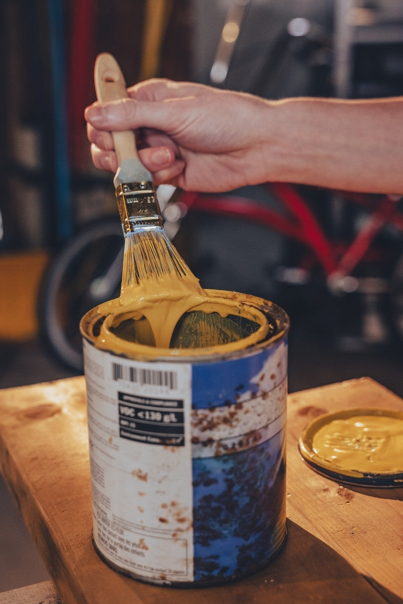 Hand holding a paintbrush over a can of yellow paint on a wooden surface.