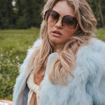 Woman wearing sunglasses and a light blue fur coat in a natural setting wearing Soul Sister since 1969