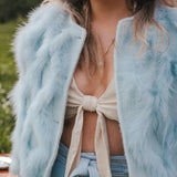 Person wearing a light blue fluffy jacket over a beige top and denim pants, with a blurred natural background wearing Soul Sister since 1969.