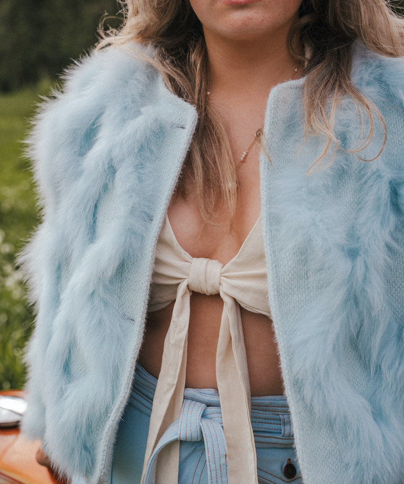 Person wearing a light blue fluffy jacket over a beige top and denim pants, with a blurred natural background wearing Soul Sister since 1969.