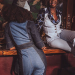 Two women wearing funky 1970s inspired denim outfits sitting at a retro bar, one in a denim jumpsuit and the other in a funky patterned outfit.