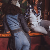 Two women wearing funky 1970s inspired denim outfits sitting at a retro bar, one in a denim jumpsuit and the other in a funky patterned outfit.