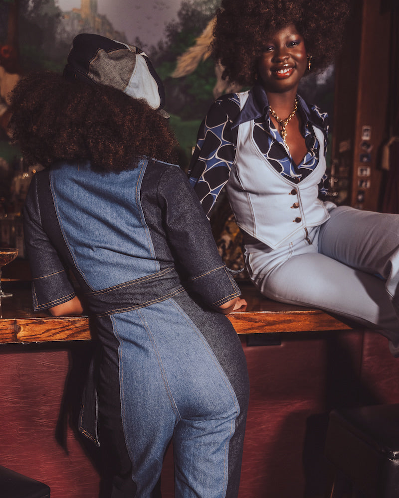 Two women sitting at a bar, one wearing a denim jumpsuit and the other in a patterned outfit.