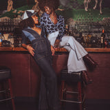 Two women wearing funky 1970s denim outfits - one is wearing a funky denim  jumpsuit, sitting at a retro bar with a mural of horses on the wall.