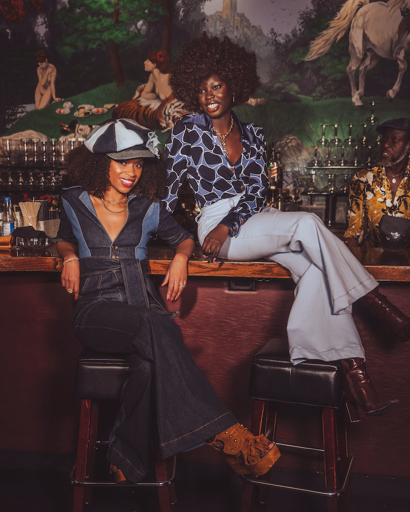 Two women wearing retro denim jumpsuits sitting at a bar with a vintage mural in the background