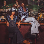 Two women wearing retro denim jumpsuits sitting at a bar with a vintage mural in the background
