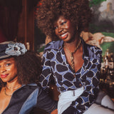 Two women sitting together, one wearing a funky 1970s inspired patterned top and the other in a denim jumpsuit, with a blurred background.