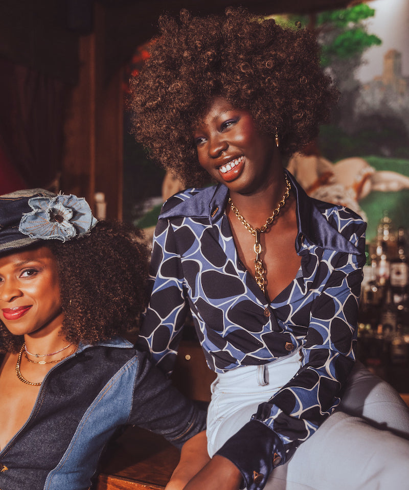 Two women sitting together, one wearing a funky 1970s inspired patterned top and the other in a denim jumpsuit, with a blurred background.