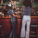 Two people standing at a retro style bar with a vintage mural in the background wearing 1970s style bellbottom jeans and a denim jumpsuit