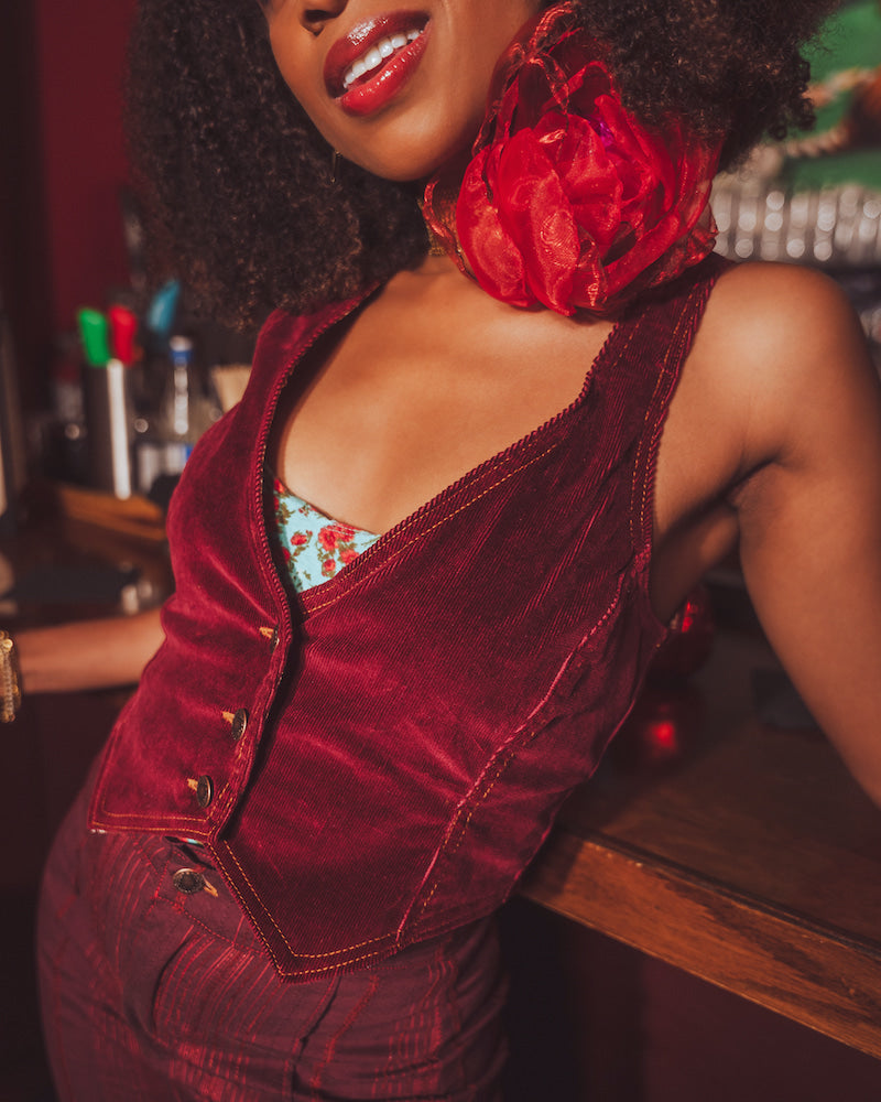 Woman wearing a red velvet 1970s outfit with a large red flower choker, standing in a retro bar setting.