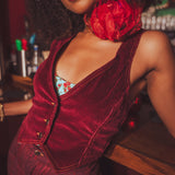 Woman wearing a red velvet 1970s outfit with a large red flower choker, standing in a retro bar setting.