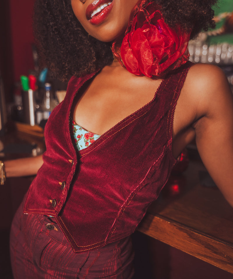 Woman wearing a red velvet 1970s outfit with a large red flower choker, standing in a retro bar setting.