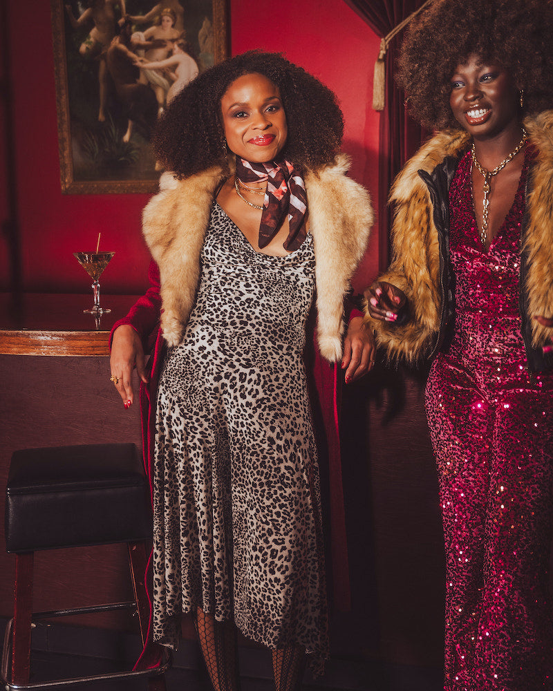 Two women in 1970s disco style outfits with fur coats in a retro bar setting.