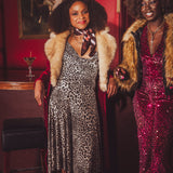 Two women in 1970s disco style outfits with fur coats in a retro bar setting.