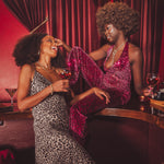 Two women in glamorous disco style outfits sitting at a 1970s speakeasy bar with drinks, surrounded by red curtains.