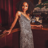 Woman in a  black and white leopard print dress standing at a 1970s style bar counter with a dark, possibly vintage interior.