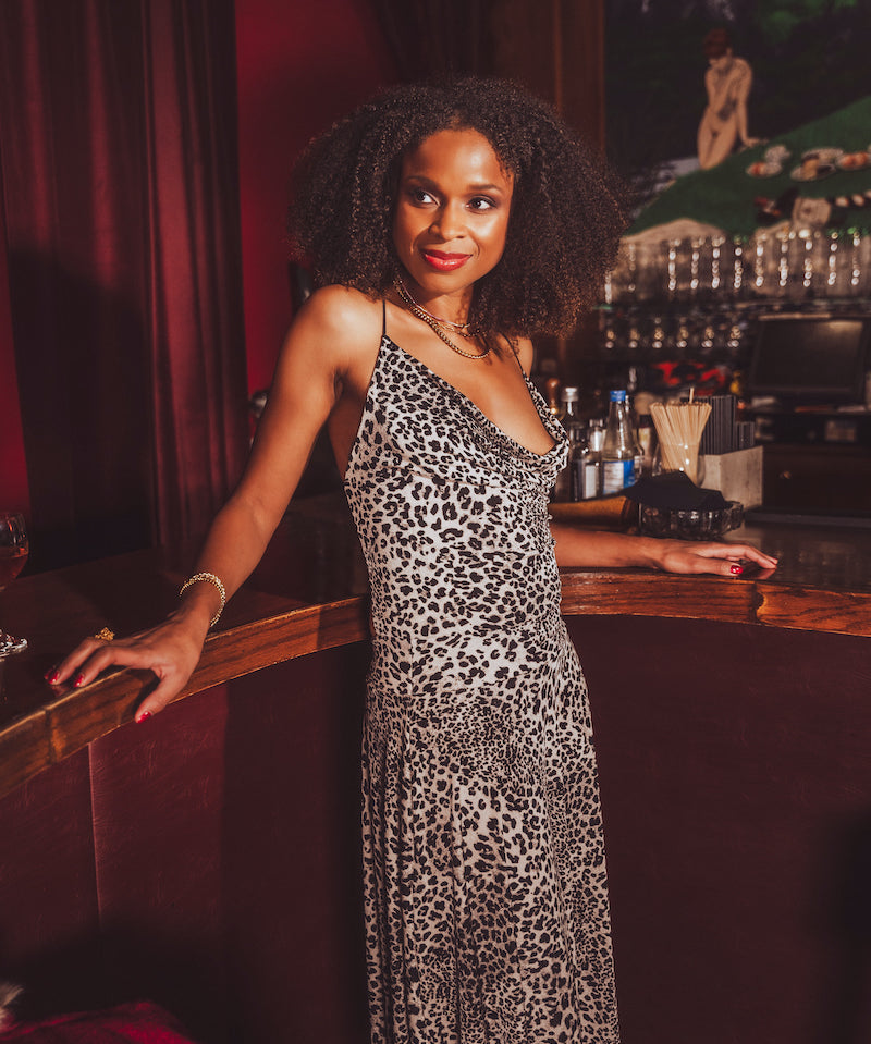 Woman in a  black and white leopard print dress standing at a 1970s style bar counter with a dark, possibly vintage interior.