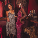 Two women in glamorous outfits standing in a bar with red curtains and bar equipment.