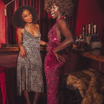Two women in glamorous 1970s retro outfits standing in a  vintage bar setting.