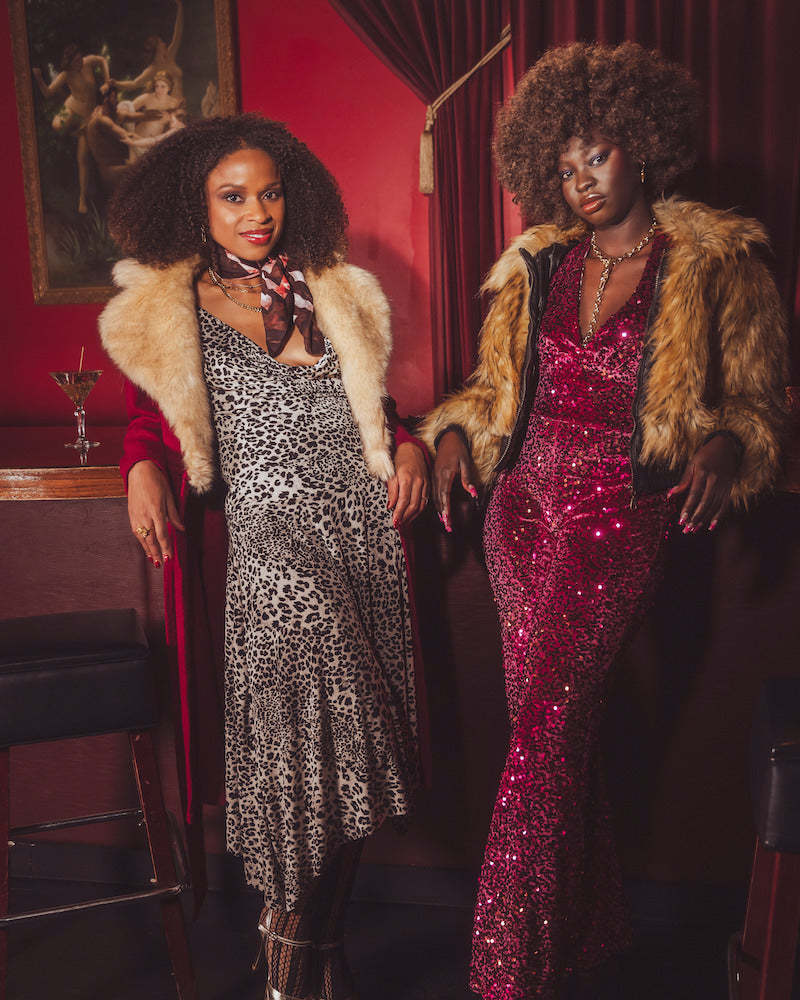 Two women in glamorous 1970s retro inspired outfits standing in a  vintage style speakeasy room with red walls and a painting.