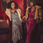 Two women in glamorous 1970s retro inspired outfits standing in a  vintage style speakeasy room with red walls and a painting.