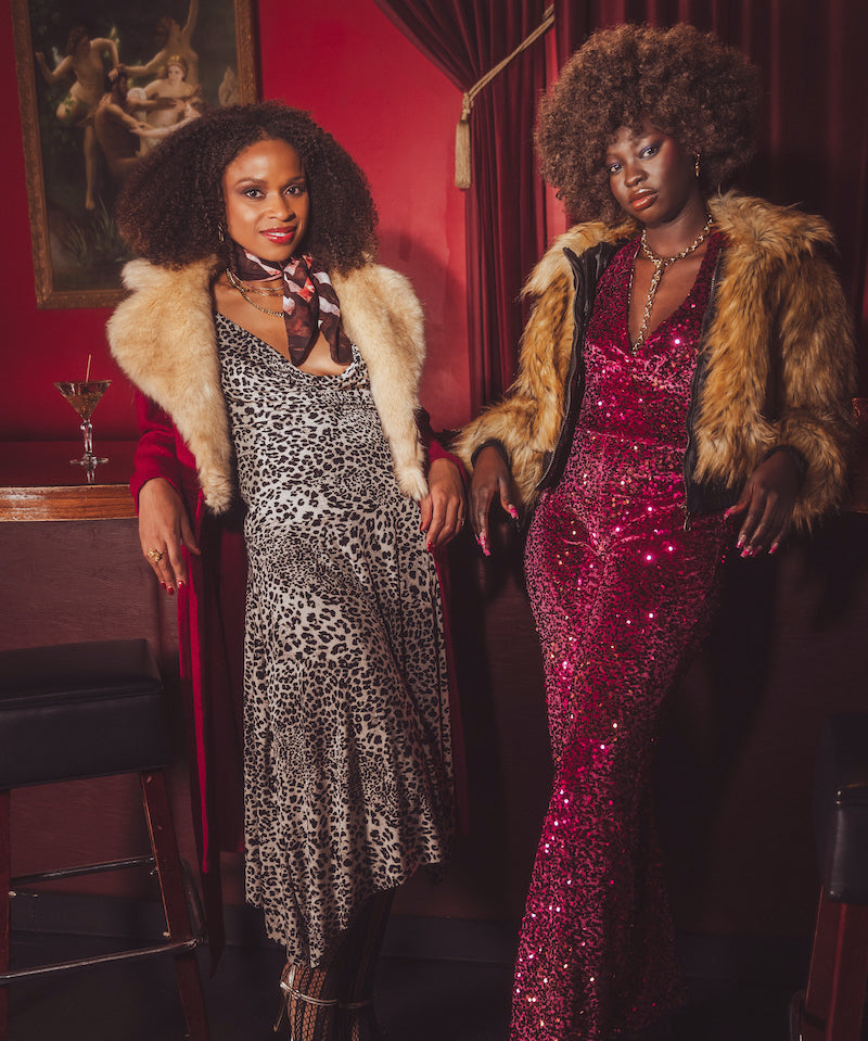 Two women in glamorous 1970s retro inspired outfits standing in a  vintage style speakeasy room with red walls and a painting.