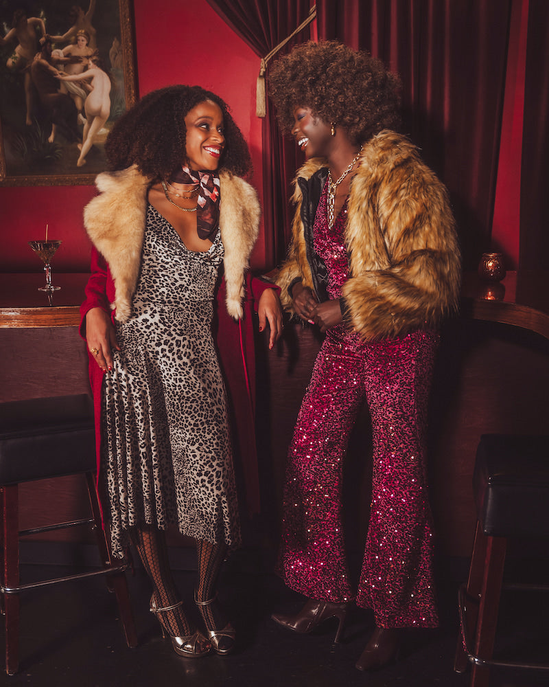 Two women in glamorous  studio 54 style outfits with fur coats standing in a r1970s style speakeasy bar