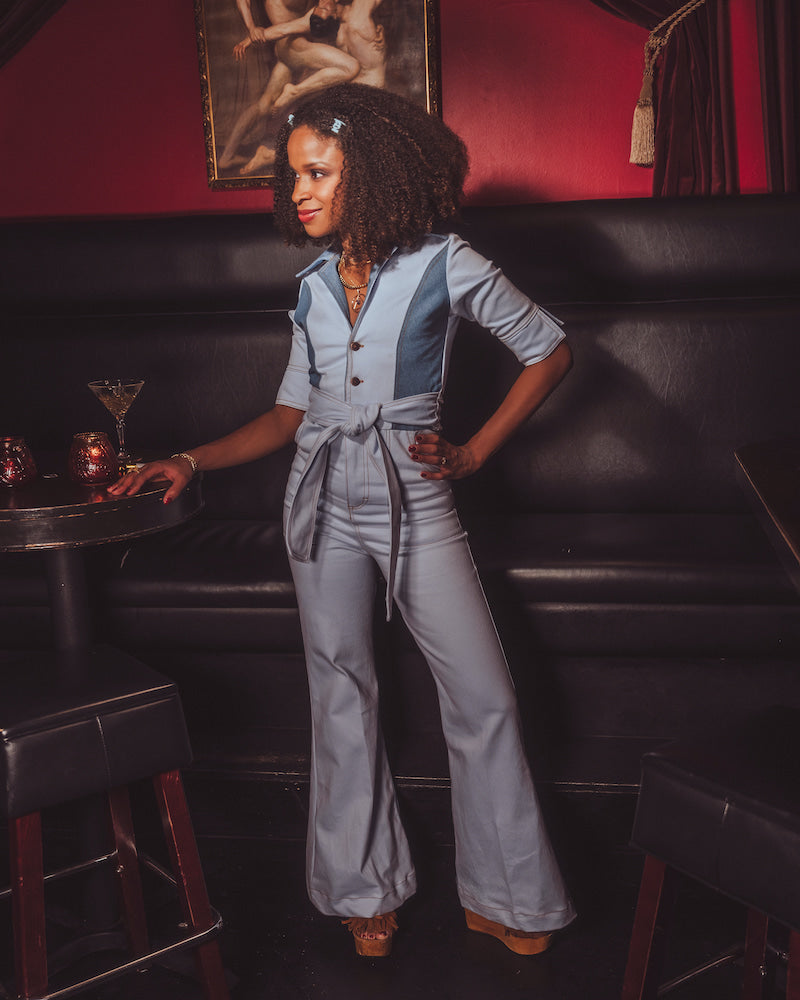 Woman in a gray jumpsuit standing in a bar with a painting on the wall.