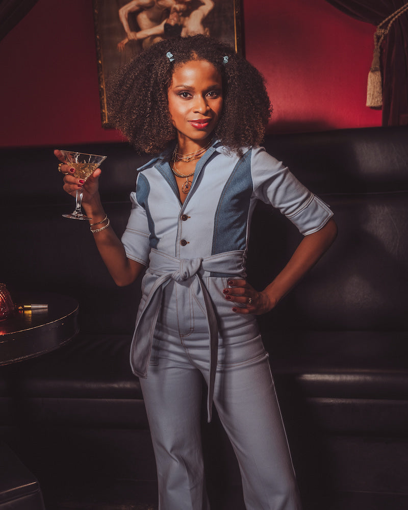 Woman in a gray jumpsuit holding a cocktail in a dimly lit room.