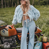 Woman in a light blue fur coat standing next to a motorcycle in a grassy field. Soul Sister since 1969.