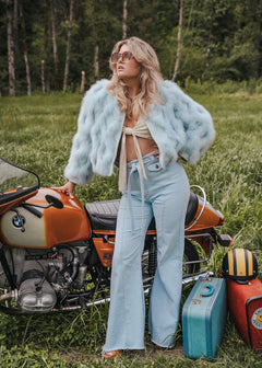Woman in a light blue fur coat standing next to a motorcycle in a grassy field. Soul Sister since 1969.