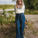 Woman in a white t-shirt and blue jeans standing next to a motorcycle in a natural setting Woman wearing a peace symbol shirt with text outdoors wearing Soul Sister since 1969.
