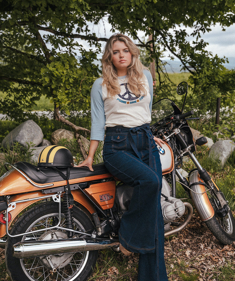 Woman sitting on a motorcycle in a natural setting with trees and rocks wearing Soul Sister since 1969..