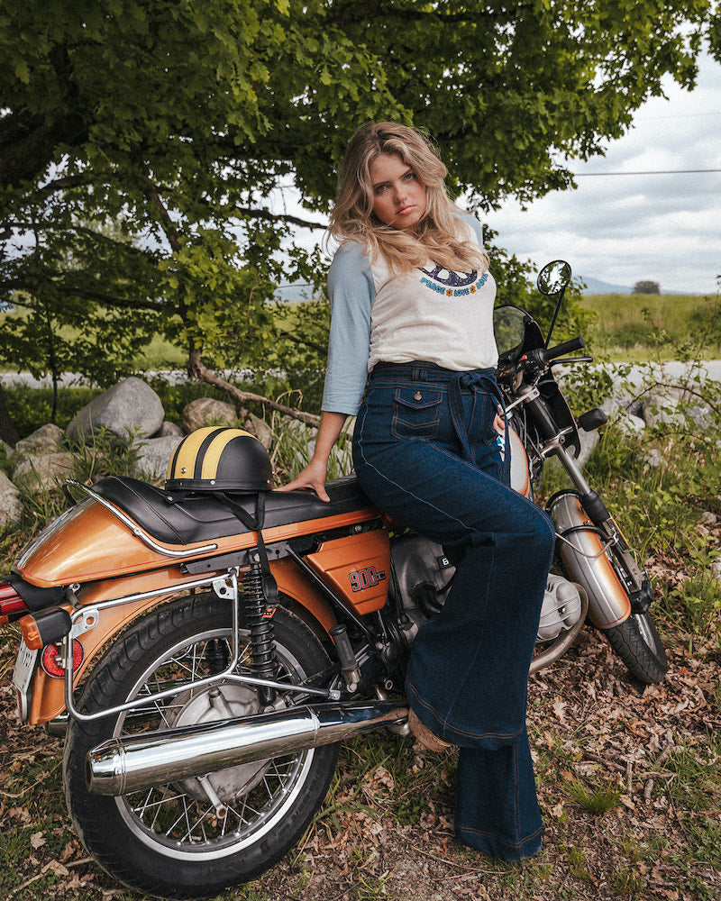 Woman sitting on a motorcycle in a natural setting with trees and grass wearing Soul Sister since 1969..