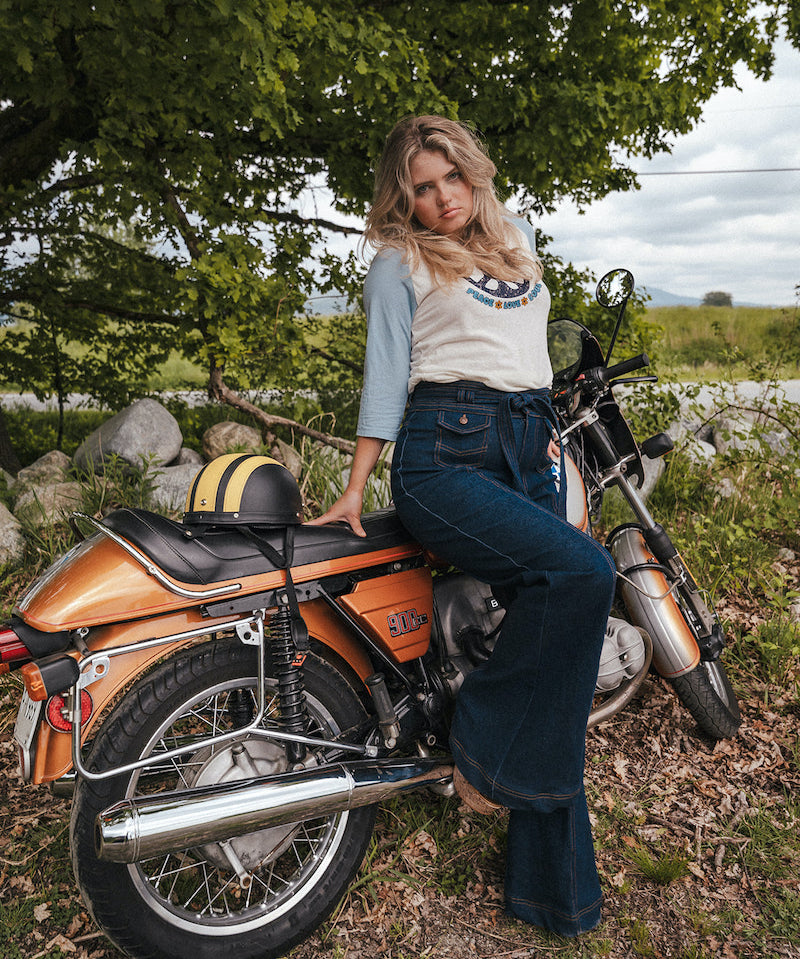 Woman sitting on a motorcycle in a natural setting with trees and grass wearing Soul Sister since 1969..