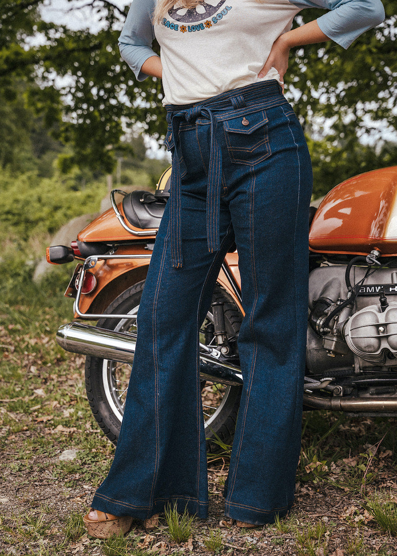 Person wearing blue jeans standing next to an orange motorcycle in a natural setting wearing Soul Sister since 1969.