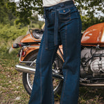 Person wearing blue jeans standing next to an orange motorcycle in a natural setting wearing Soul Sister since 1969.