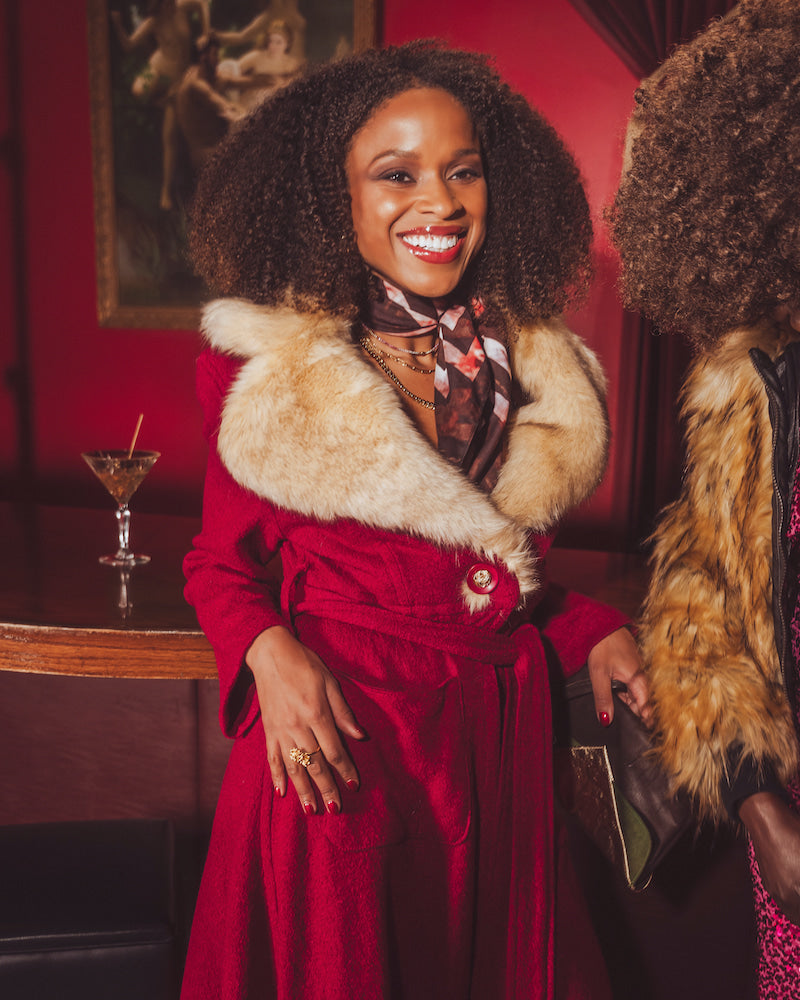 Woman in a red vintage inspired coat with fur collar standing in a 1970s style bar with a red wall and a drink in the background.