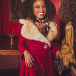 Woman in a red vintage inspired coat with fur collar standing in a 1970s style bar with a red wall and a drink in the background.