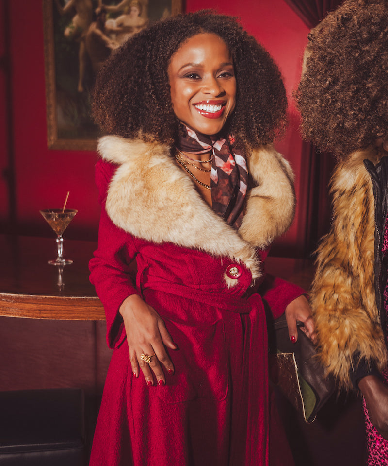 Woman in a red vintage inspired coat with fur collar standing in a 1970s style bar with a red wall and a drink in the background.