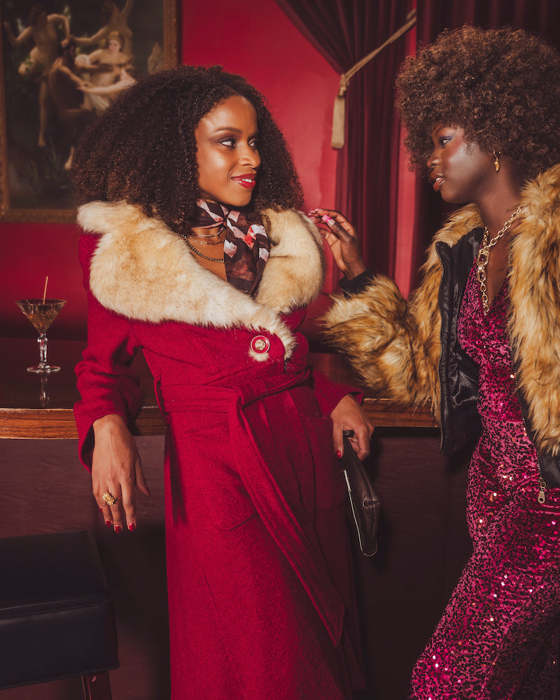 Two women in glamorous 70s disco inspired outfits with fur collars in a retro vintage setting.