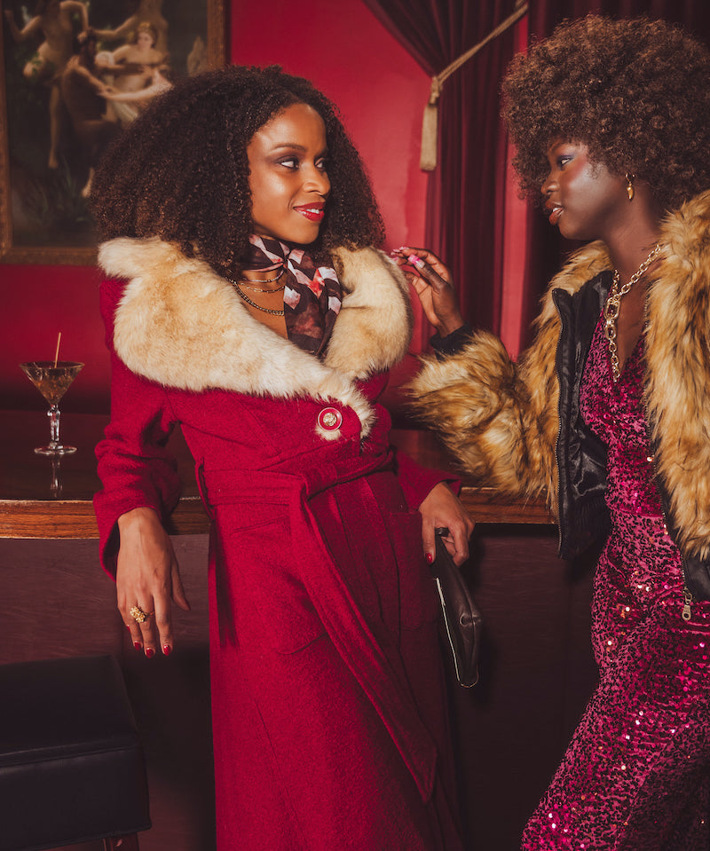 Two women in glamorous 70s disco inspired outfits with fur collars in a retro vintage setting.