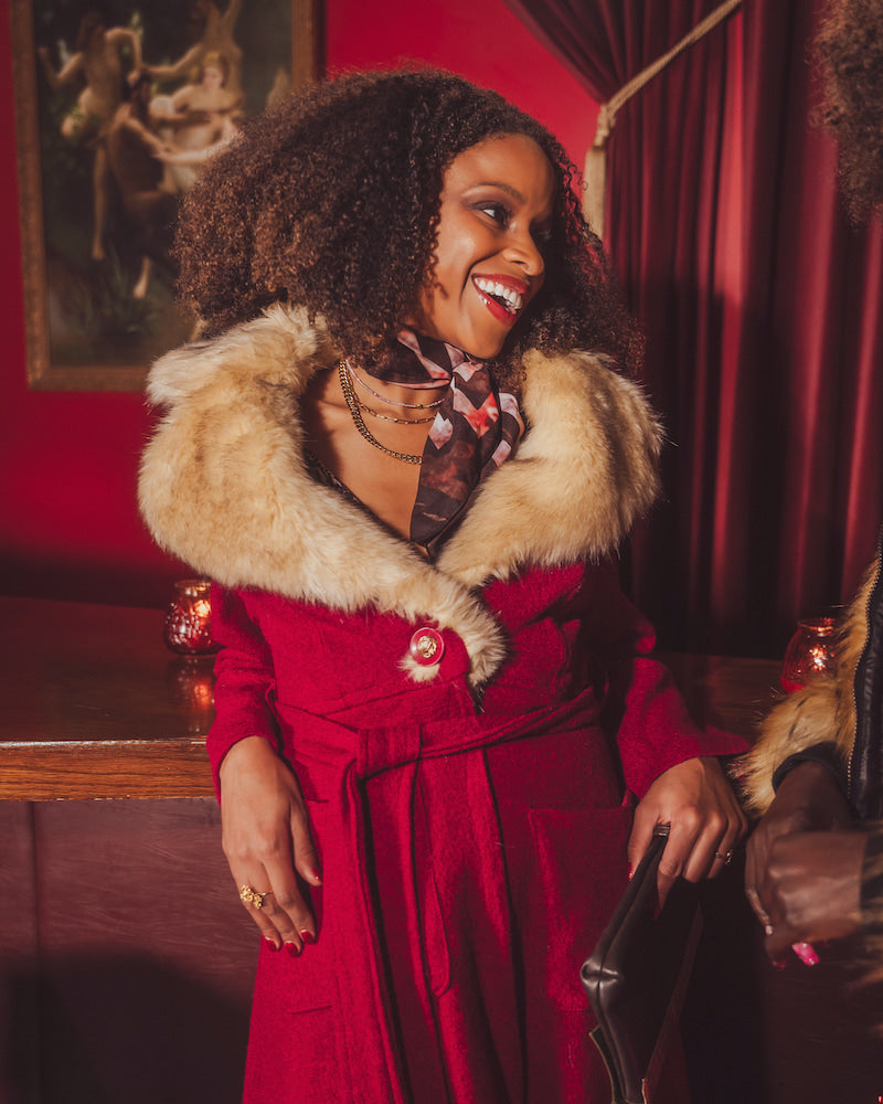 Woman in a red 1970s inspired coat with fur collar standing in a retro speakeasy bar  with red walls and curtains.