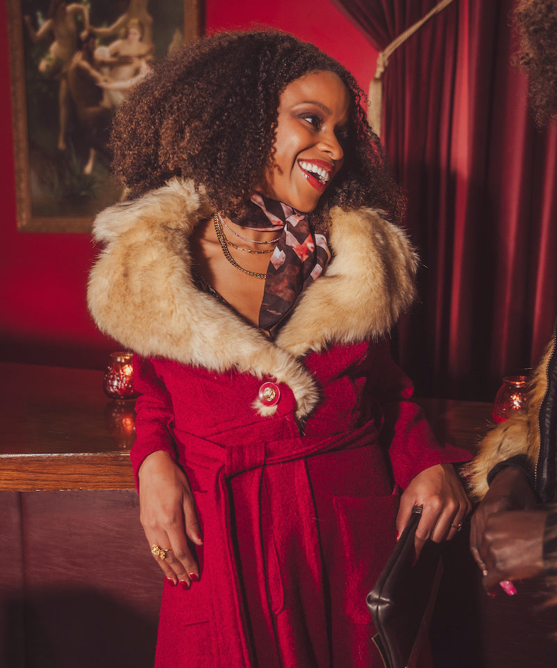 Woman in a red 1970s inspired coat with fur collar standing in a retro speakeasy bar  with red walls and curtains.