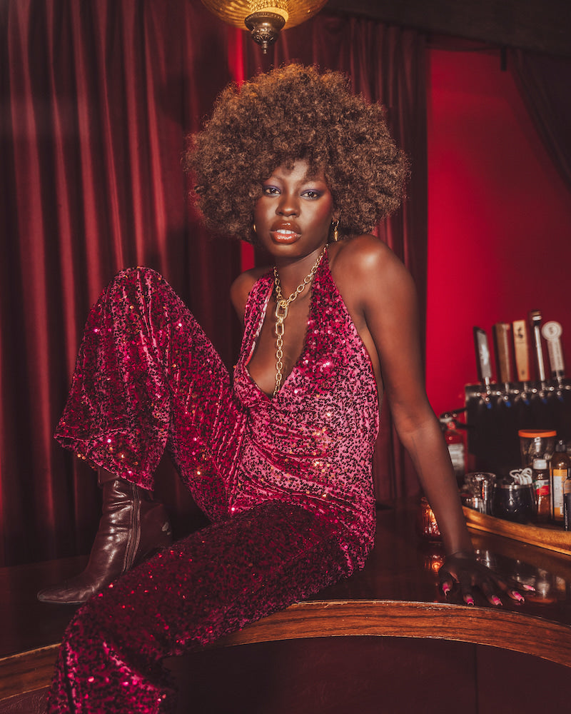 Woman in a red sequin outfit sitting at a bar with red curtains and bottles in the background