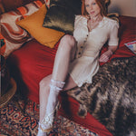 Woman in a white dress and knee-high boots sitting on a red couch with a cat.