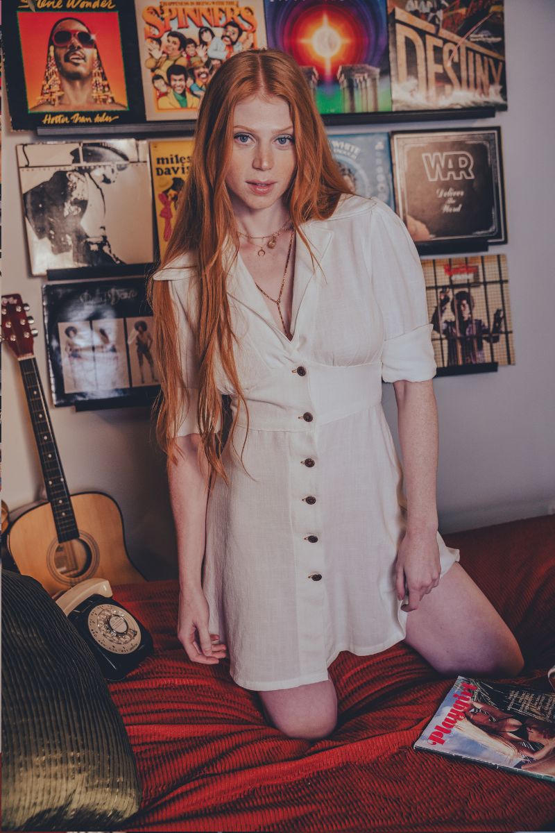 Woman in a white dress standing in a room with vinyl records and a guitar.