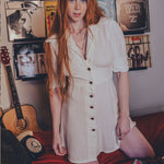 Woman in a white dress standing in a room with vinyl records and a guitar.
